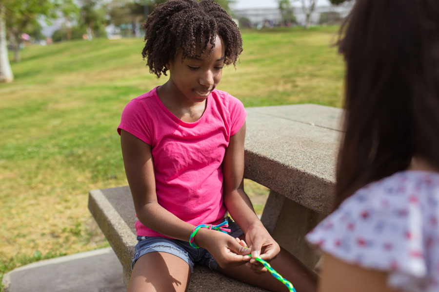 Two girls braiding a friendship bracelet on a park picnic table